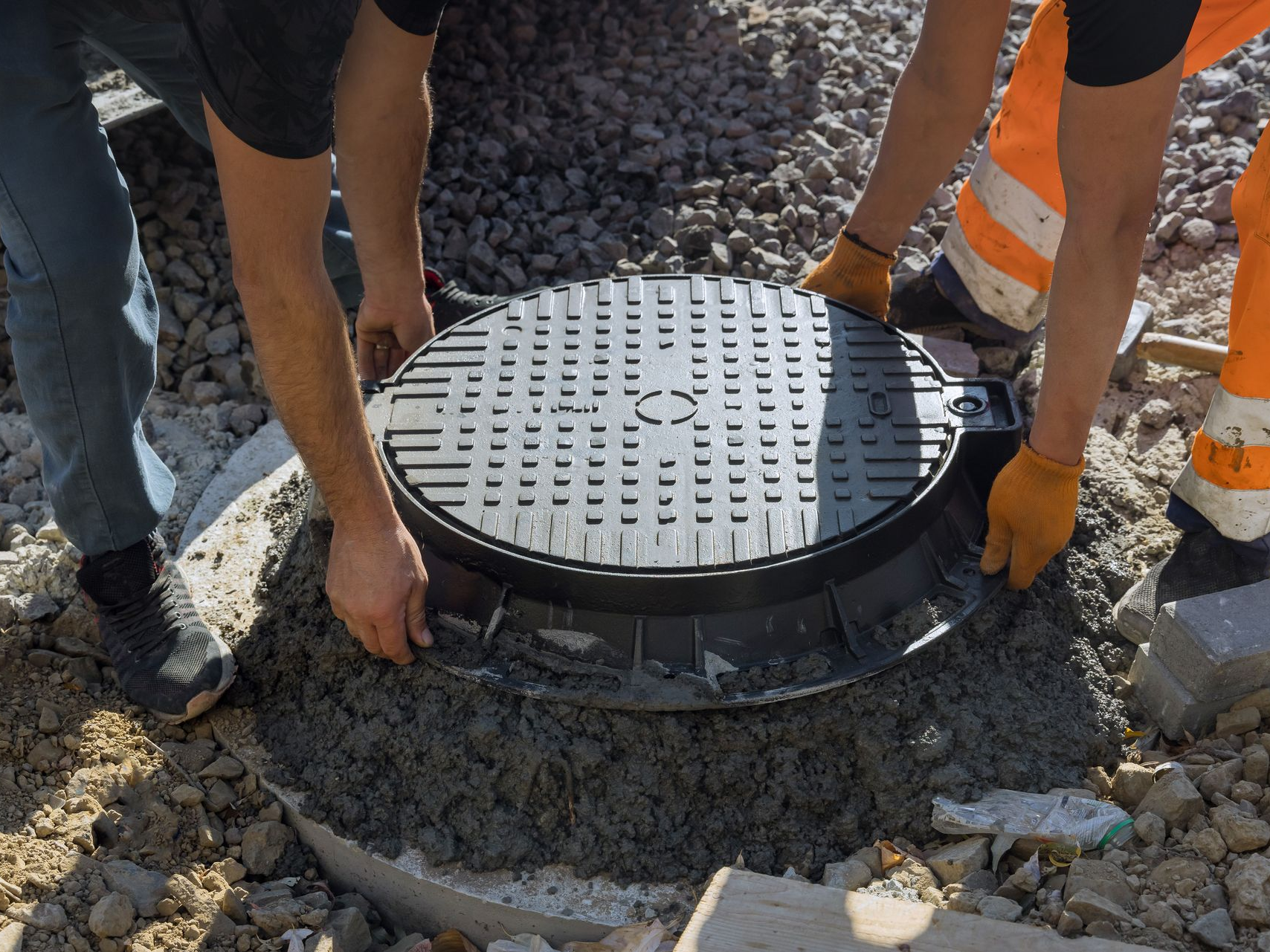 Deux personnes installent une plaque d'égout sur une base en béton. Les ouvriers portent des gants et des pantalons orange.