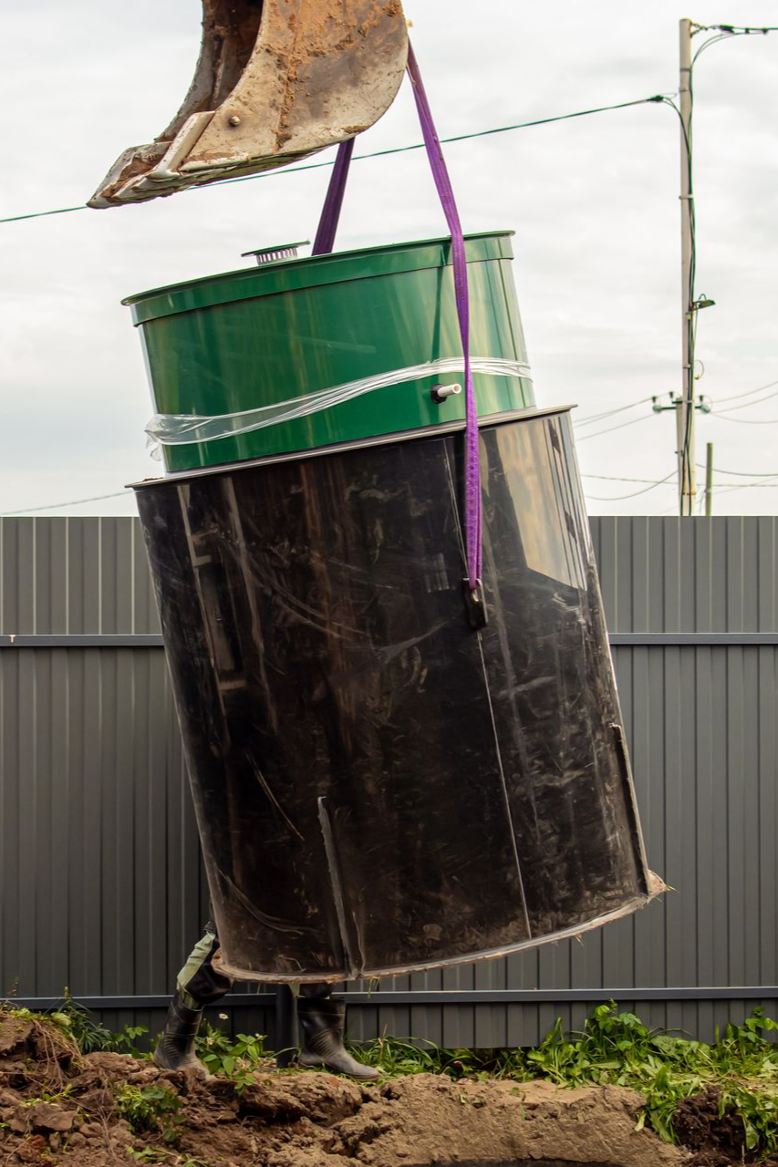 Une machine soulève un fût vert au-dessus d'un autre fût noir et de terre. Une clôture grise se dresse en arrière-plan.