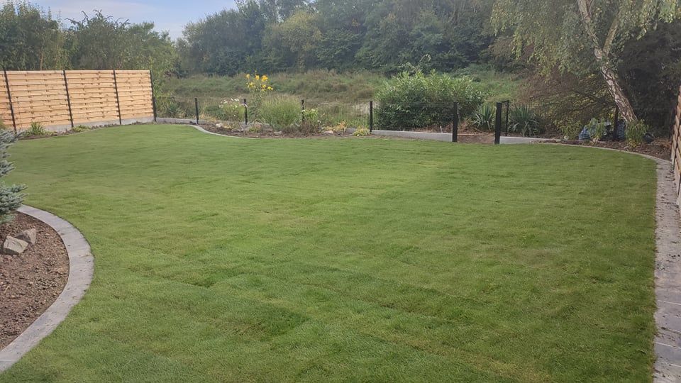 Pelouse verte dans un jardin, bordée d'un chemin de pierre et d'une clôture en bois, avec des arbres et de la verdure en arrière-plan.