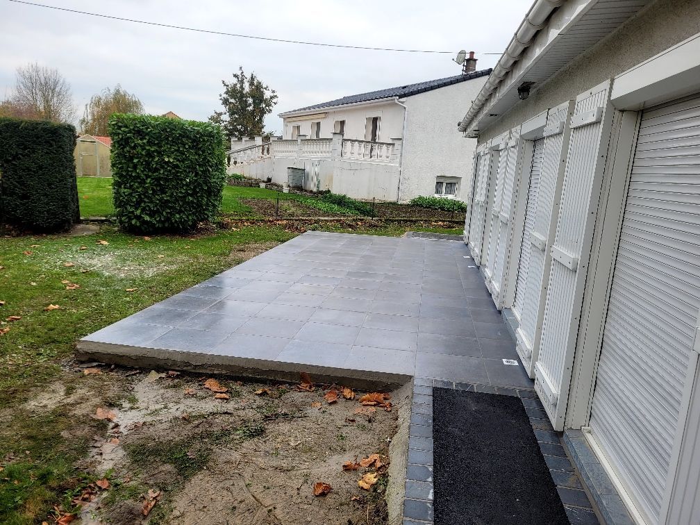 Terrasse carrelée de gris foncé jouxtant une maison blanche aux volets fermés.