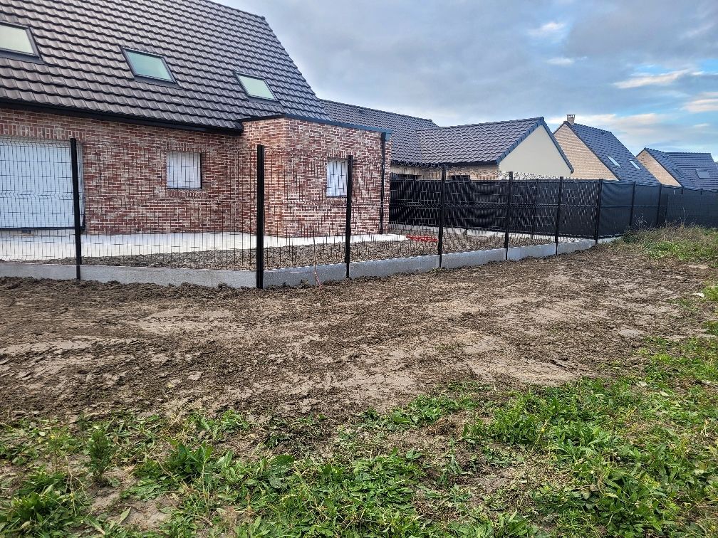 Clôture noire devant une maison en briques, socle en béton, premier plan de terre et d'herbe, ciel nuageux.