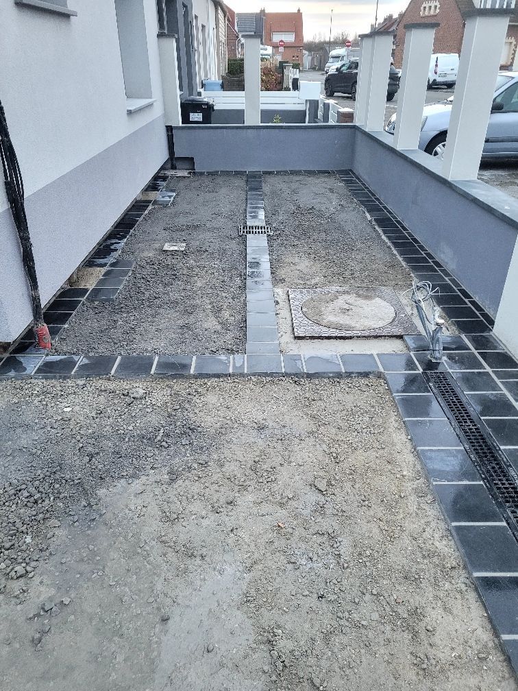 Une allée de gravier bordée de carreaux sombres, un mur de soutènement gris et un aperçu d'une rue.