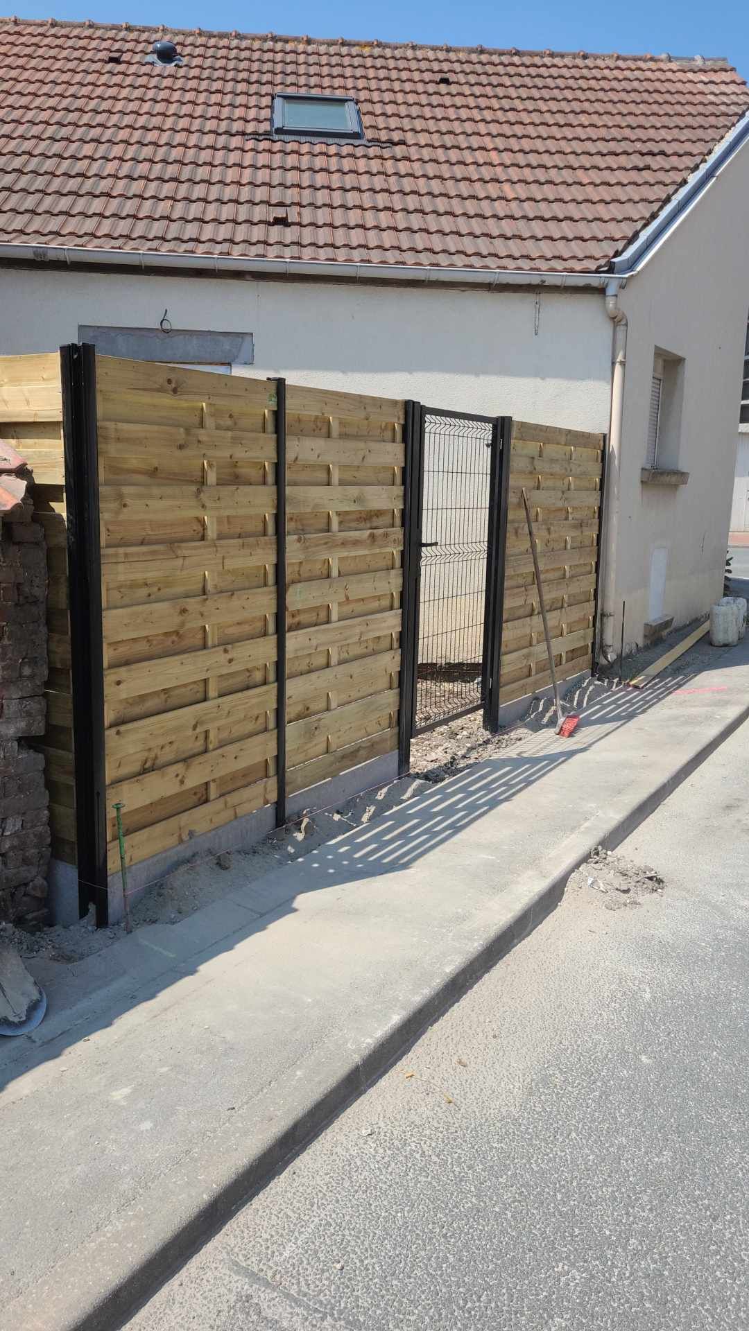Une clôture en bois avec un portail longe une route pavée, à côté d'un bâtiment au toit de tuiles.