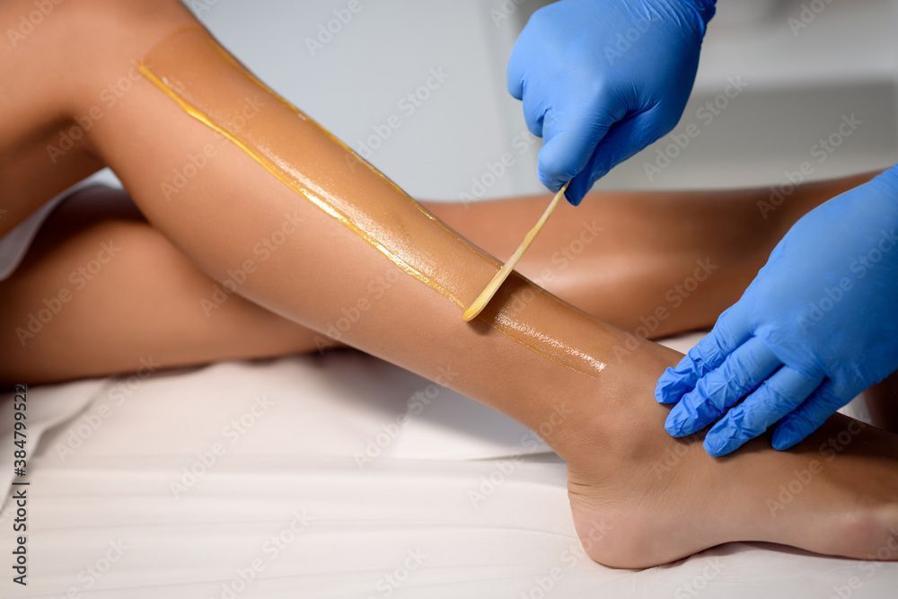 Eine Person enthaart ihr Bein. Eine behandschuhte Hand trägt das Wachs mit einem Holzspatel auf das Bein auf.