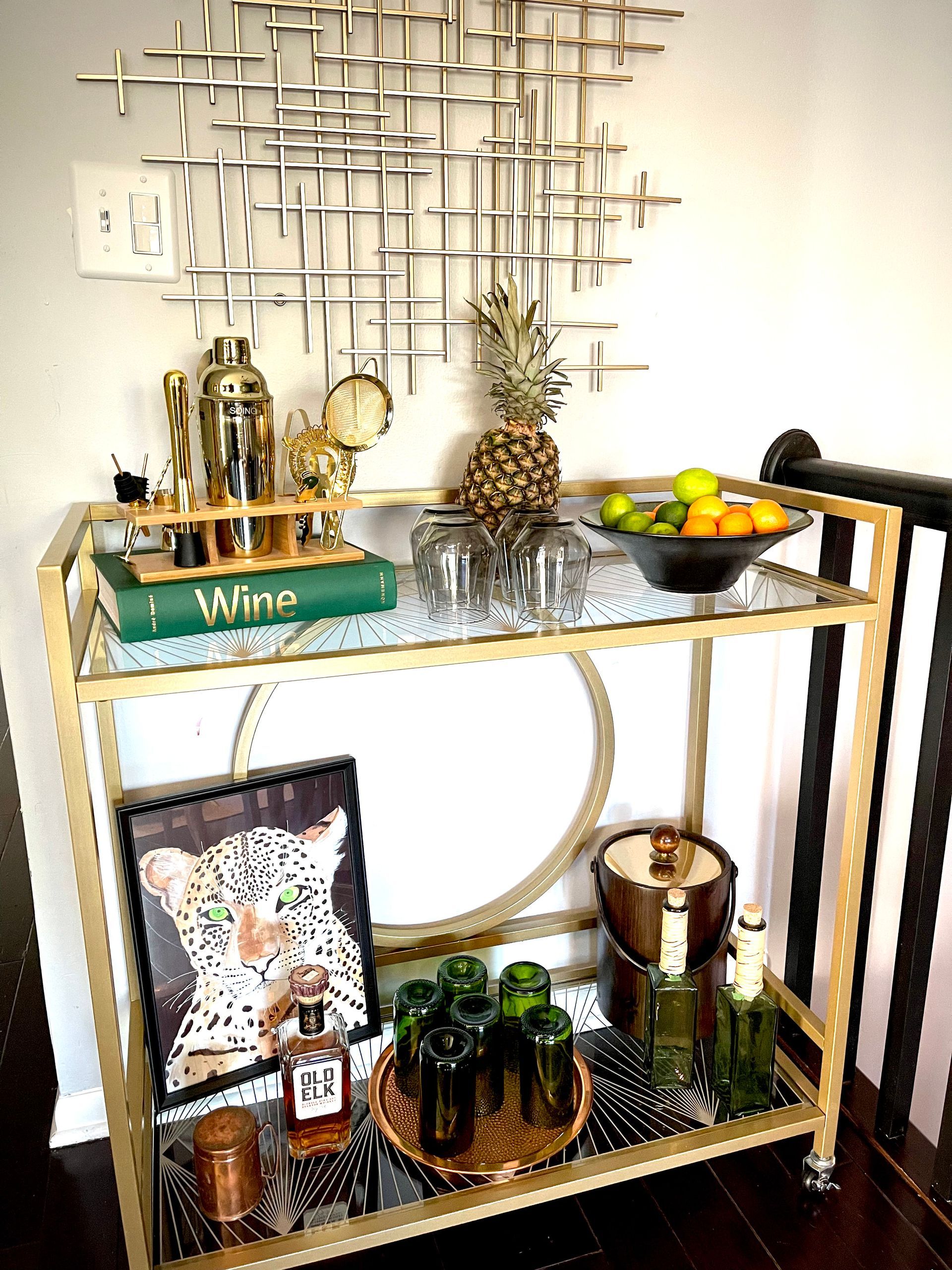 A bar cart filled with bottles , glasses , fruit , and a picture of a leopard.