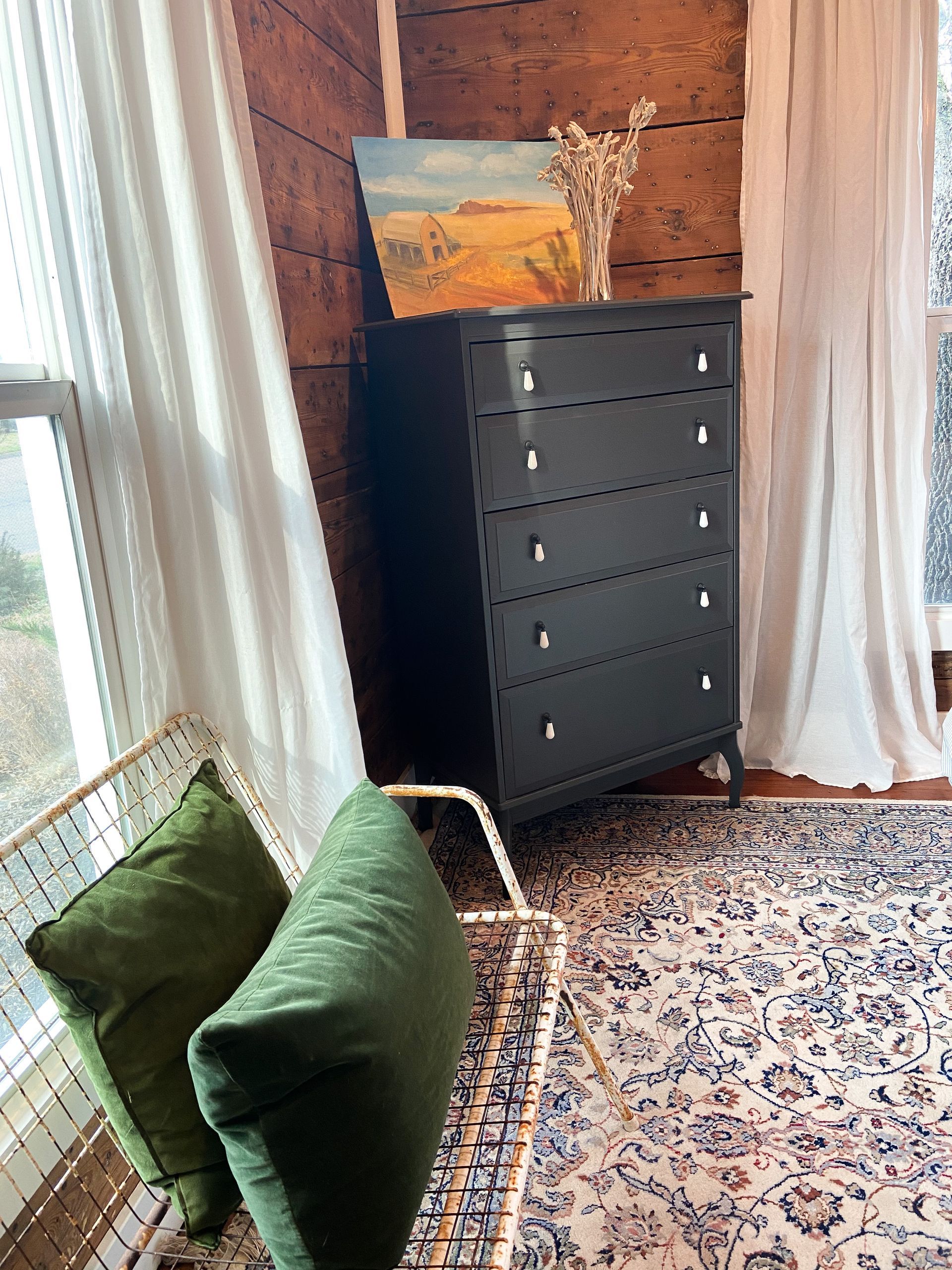 A bedroom with a black dresser , green pillows , a rug and a painting on the wall.