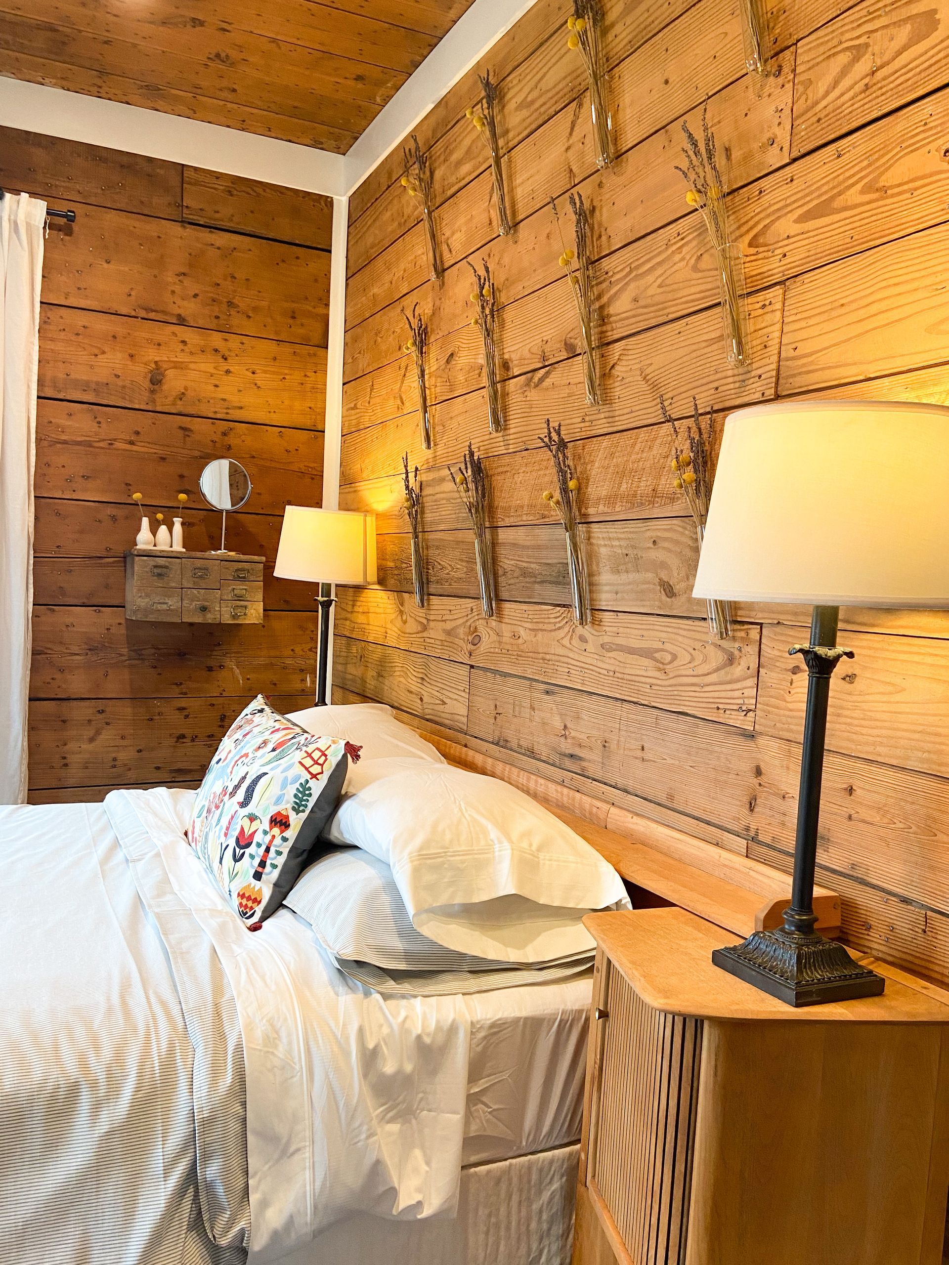 A bedroom with a bed , nightstand , lamp and wooden walls.