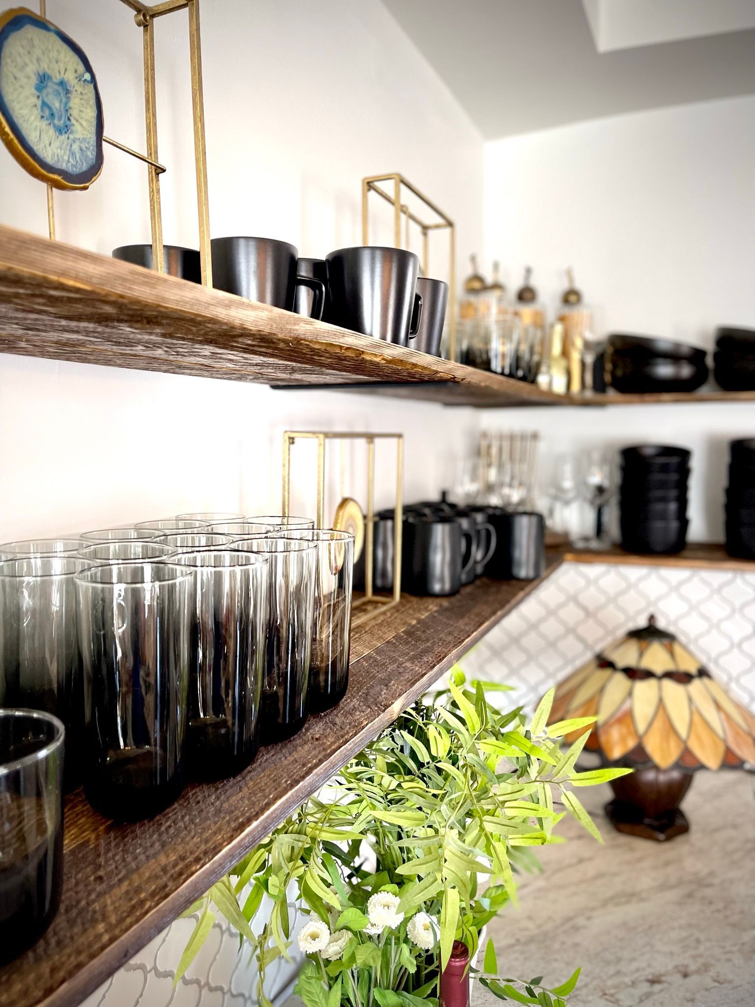 A bunch of glasses are sitting on a wooden shelf in a kitchen.