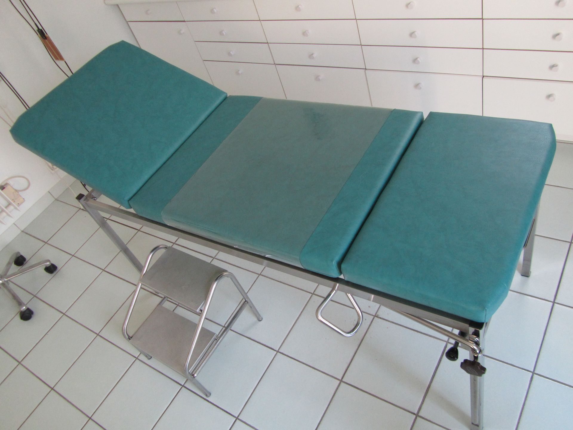 Table d'examen avec rembourrage bleu sarcelle et structure en métal, à côté d'un petit marchepied en métal.