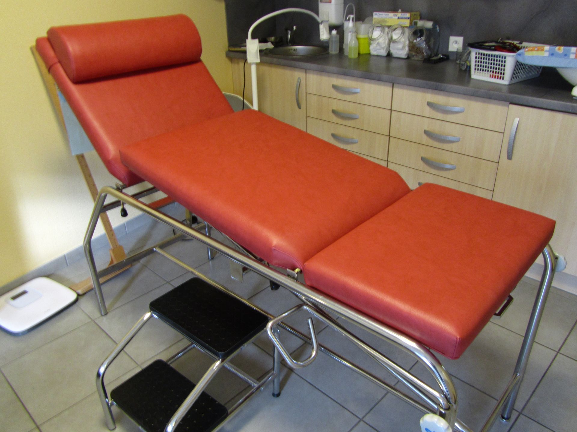Table d'examen rouge dans un cabinet médical. Comprends des sections réglables, une structure en métal et un escabeau.