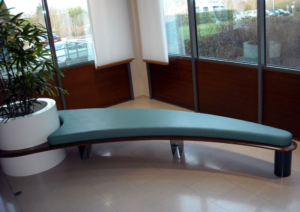 Banc courbé avec coussin bleu sarcelle, à côté d'une plante et de grandes fenêtres avec garniture en bois.