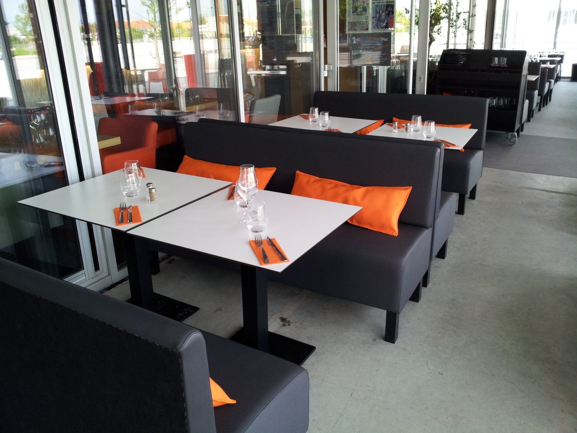 Restaurant avec tables blanches, banquettes grises et coussins orange. On aperçoit des baies vitrées et un piano.