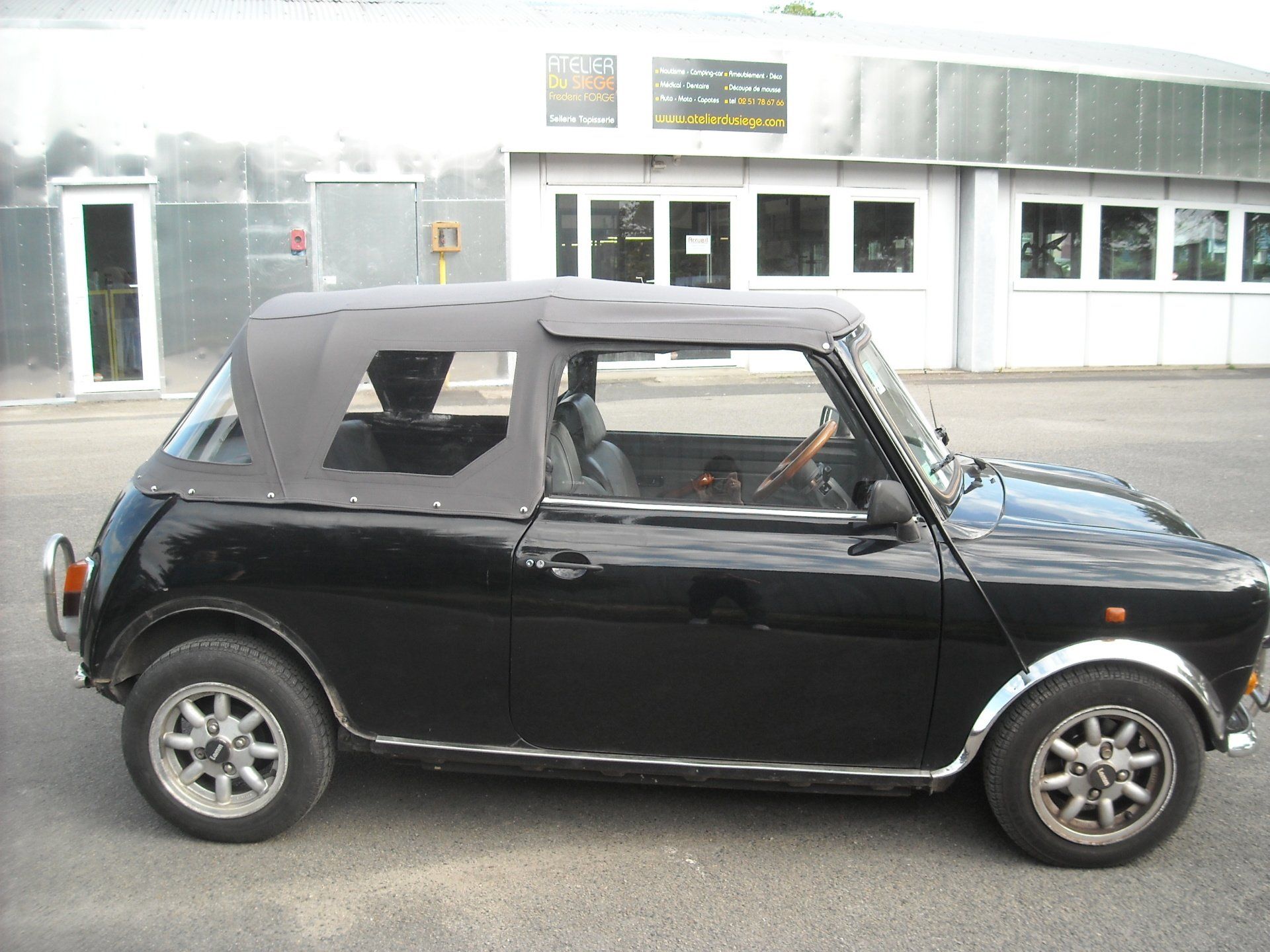 Une Mini Cooper décapotable noire est garée devant un immeuble aux nombreuses fenêtres.