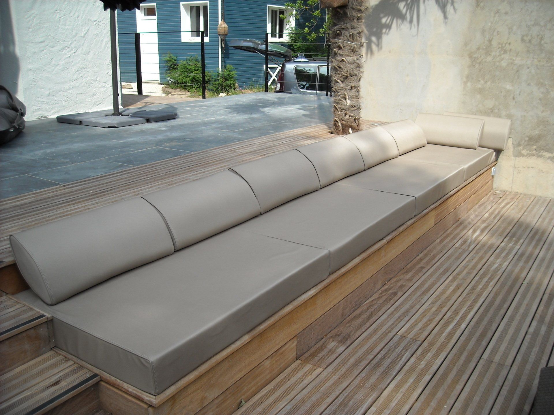 Salon d'extérieur sur une terrasse en bois, avec coussins et dossiers beiges, près d'un palmier.