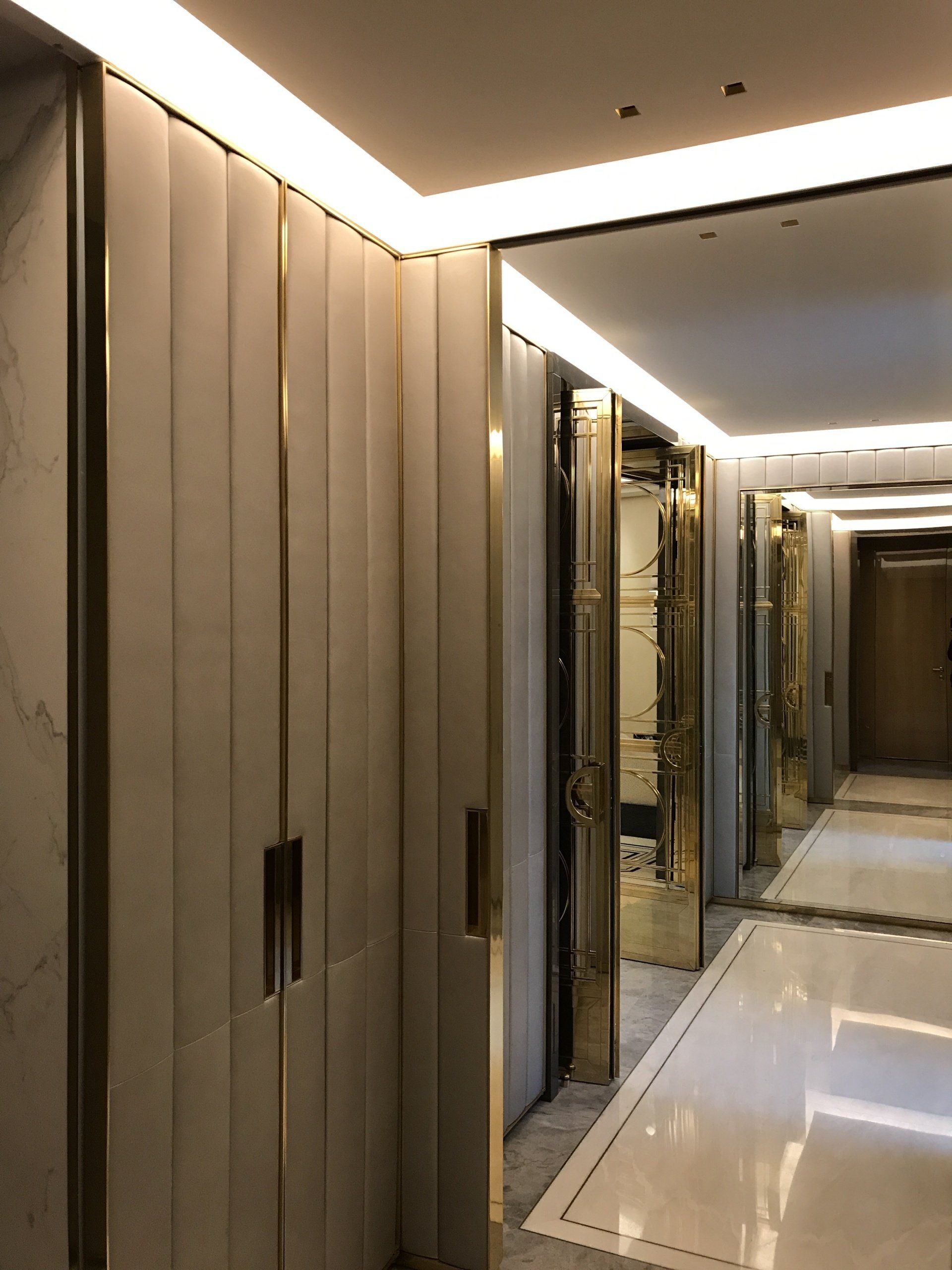 Couloir avec lambris beiges verticaux, finitions dorées et éclairage encastré. Des portes miroirs reflètent l'espace.