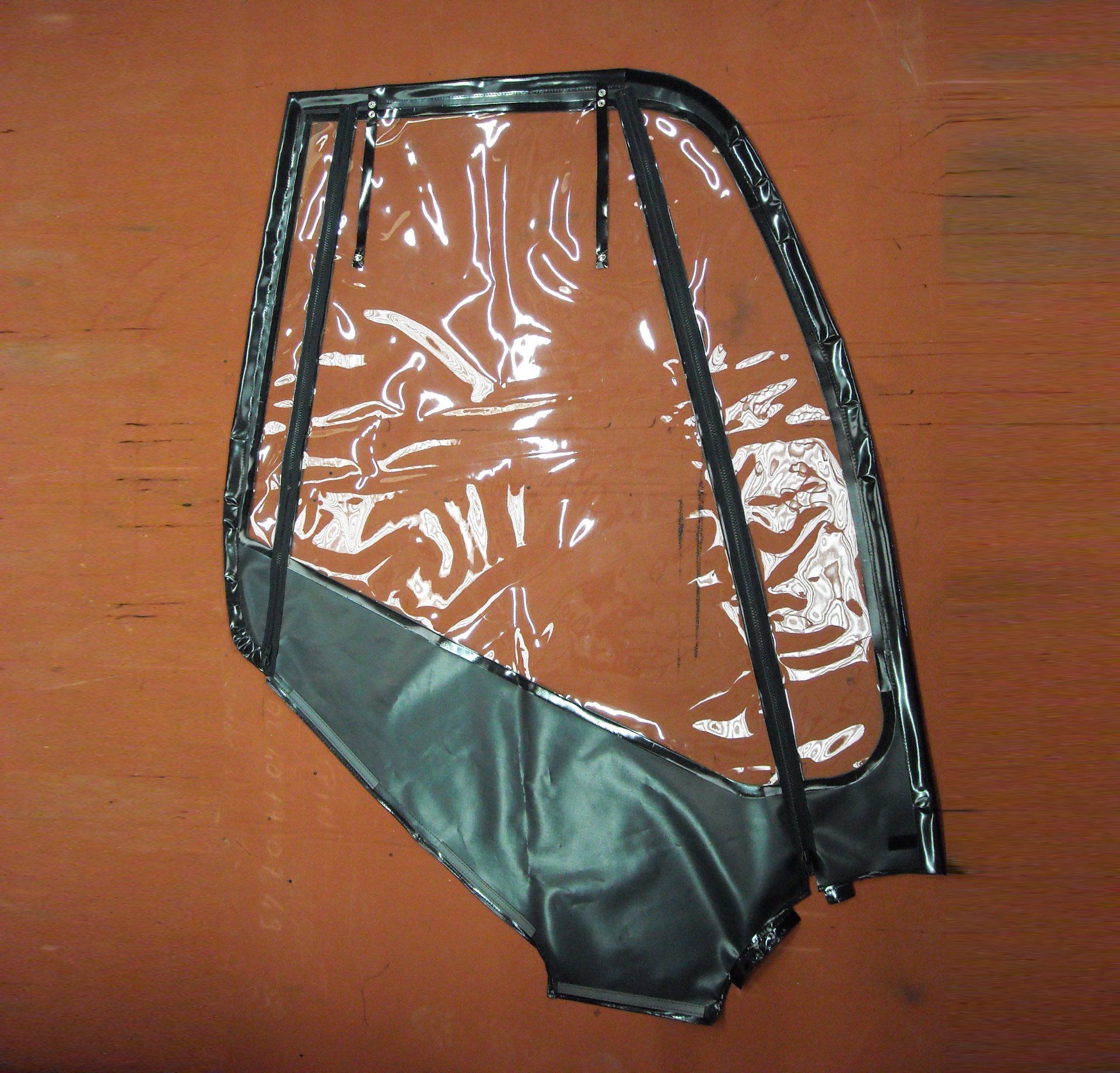 Panneau de porte de véhicule en plastique transparent avec cadre noir et partie inférieure noire, posé sur une surface orange.