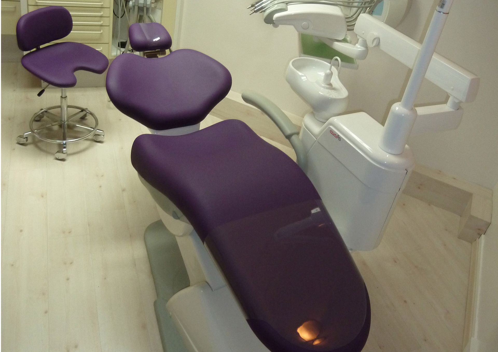 Chaise dentaire violette et tabouret dans une pièce aux murs blancs avec du matériel dentaire.