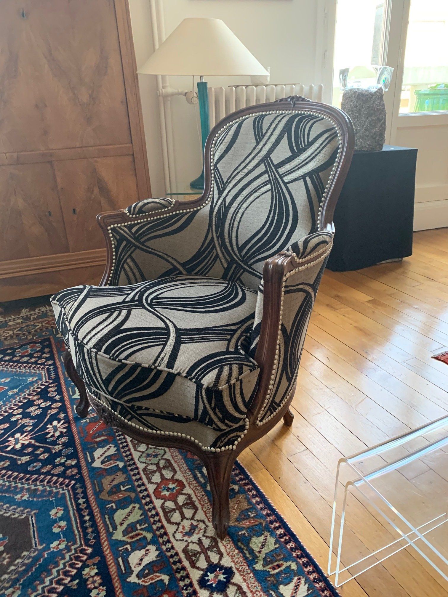 Fauteuil recouvert d'un tissu à motifs noirs et blancs, structure en bois, posé sur un tapis, près d'une armoire en bois.