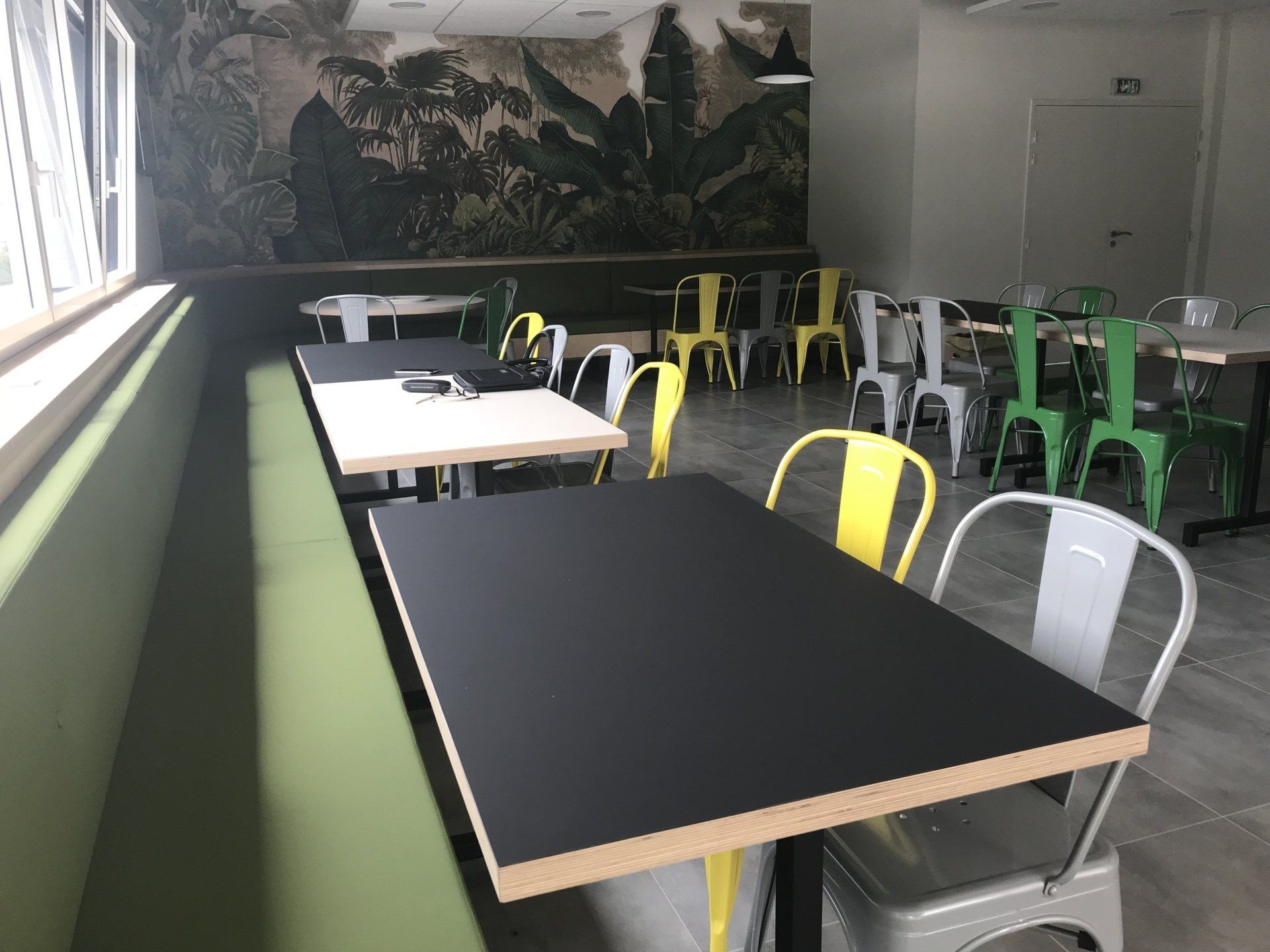 Cantine avec des tables noires, des chaises jaunes, vertes et grises, un banc vert et une fresque de la jungle sur le mur.