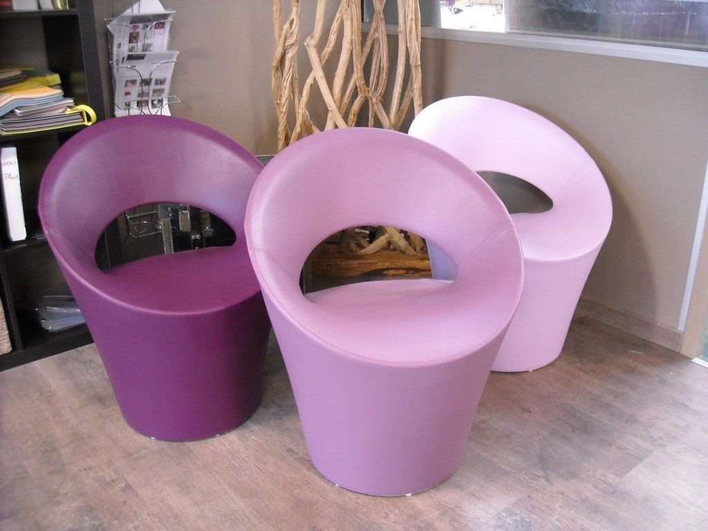 Trois chaises modernes et arrondies aux tons violets. Placées à l'intérieur, sur un parquet près d'une fenêtre.