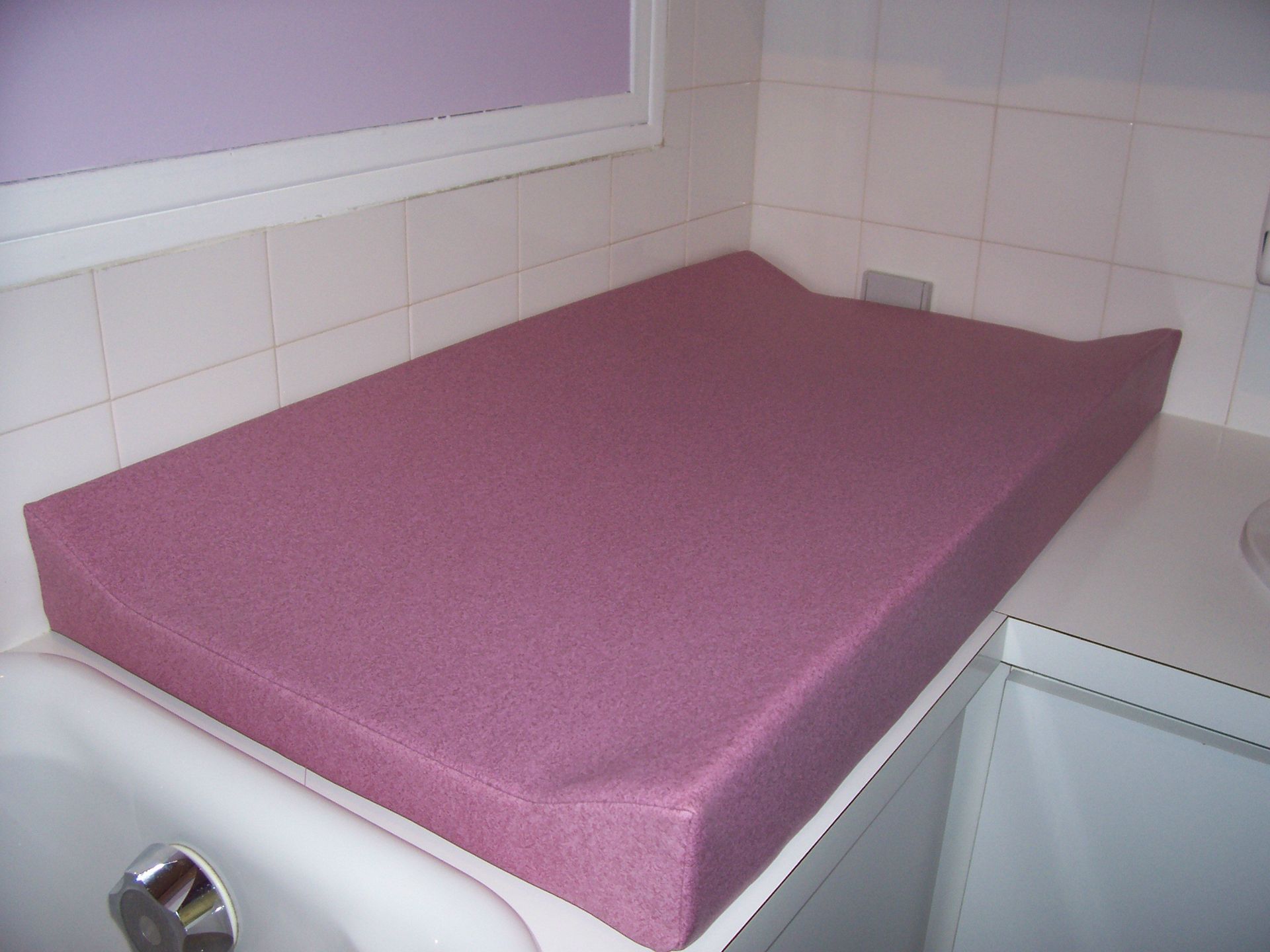 Table à langer rose sur un comptoir de salle de bains, avec une fenêtre et du carrelage en arrière-plan.