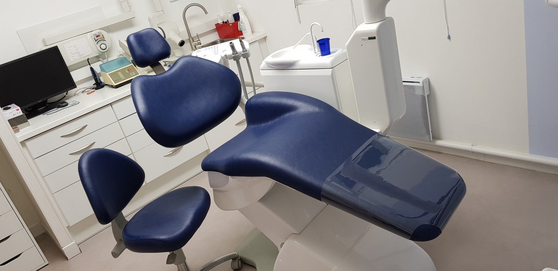 Un cabinet dentaire avec des chaises bleues, des armoires blanches et un moniteur.