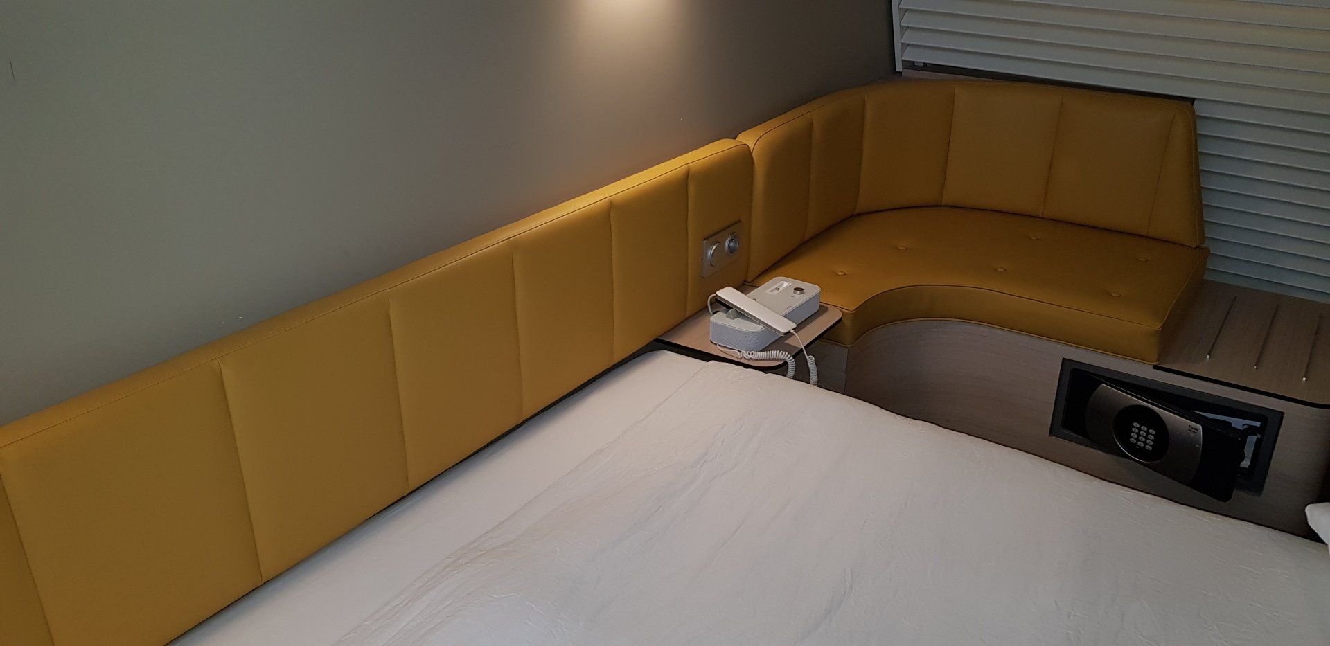 Une chambre d'hôtel avec une tête de lit jaune moutarde et un lit blanc. Une petite table avec un téléphone se trouve entre la tête de lit et le lit.