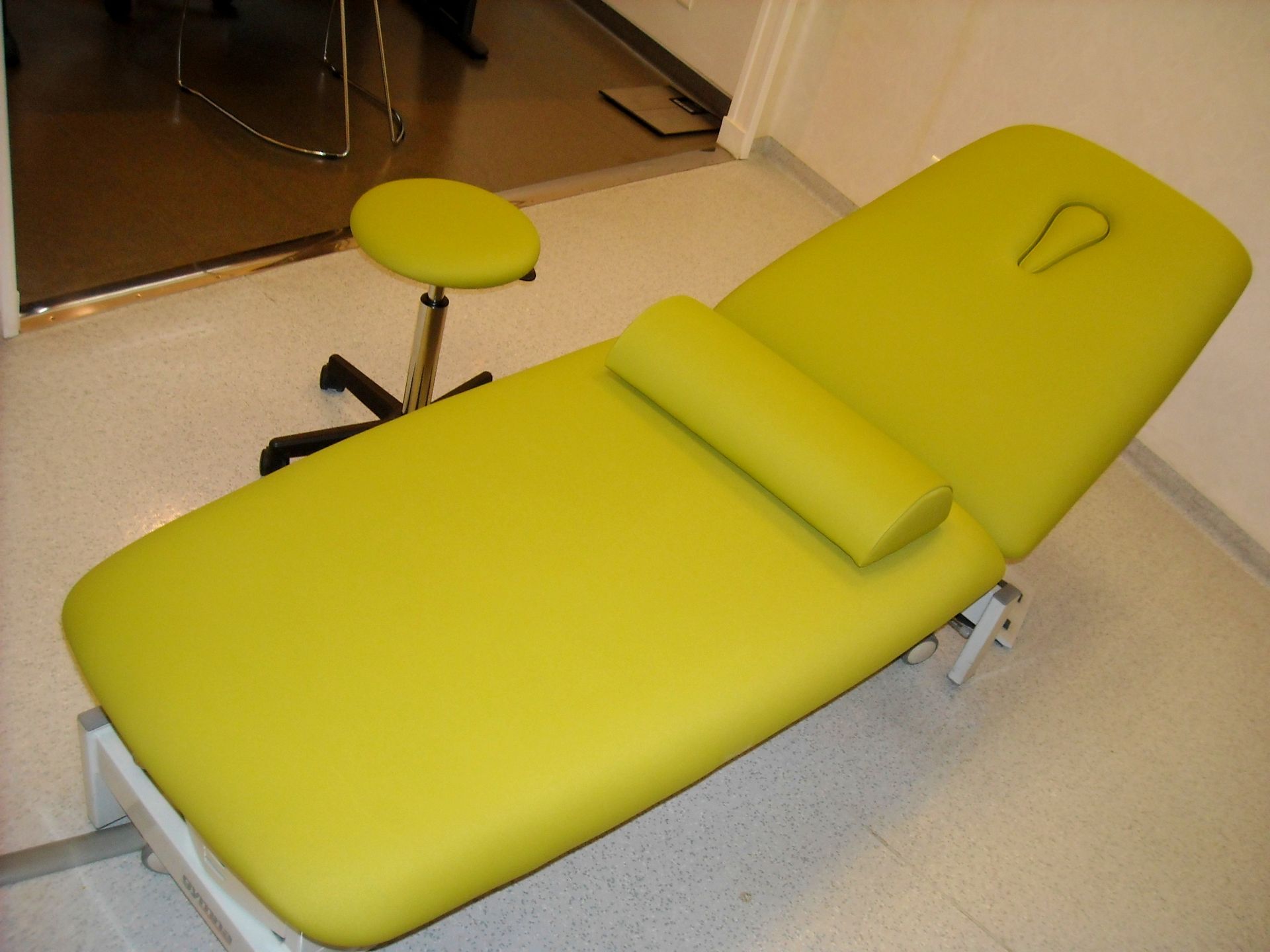 Table de massage verte avec repose-tête et appui-tête cylindrique, à côté d'un tabouret.