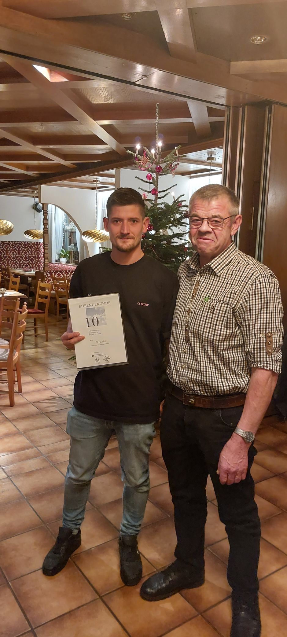 Zwei Männer stehen in einem Restaurant, einer hält eine Zeitung in der Hand. Der jüngere Mann trägt ein schwarzes Hemd und Jeans, der ältere ein gemustertes Hemd und eine schwarze Hose.