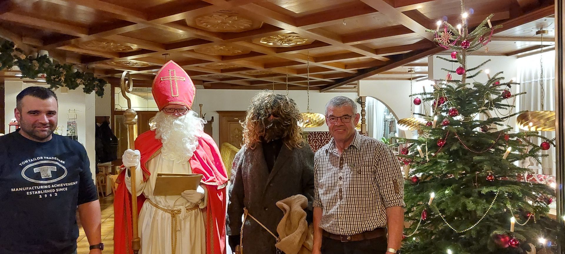Der heilige Nikolaus mit Menschen an einem Weihnachtsbaum in einem holzgetäfelten Raum.