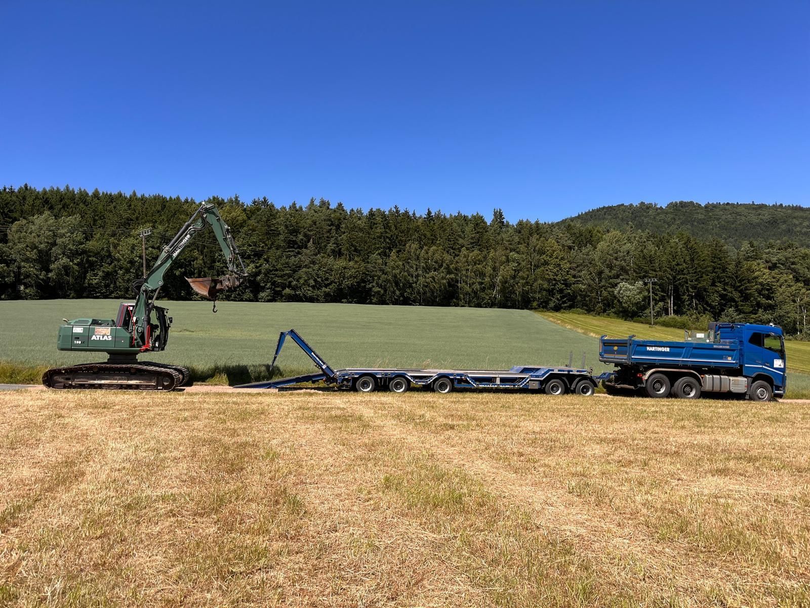 Ein grüner Bagger lädt Material auf einen Pritschenanhänger, der von einem blauen Lastwagen auf einem Feld gezogen wird.