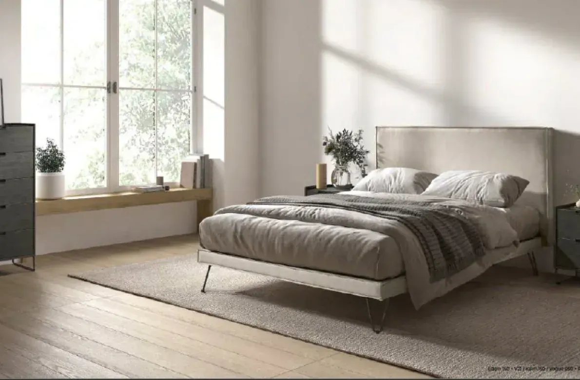 Dormitorio moderno con cama gris, ventana grande, cómoda y luz natural suave.