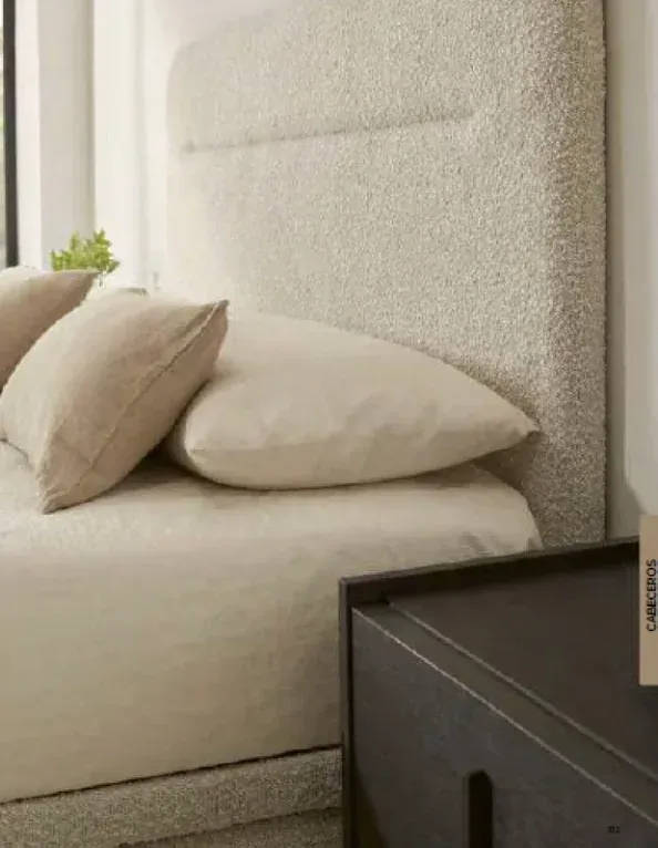Cama beige con almohadas junto a una mesita de noche negra en un dormitorio con iluminación tenue.