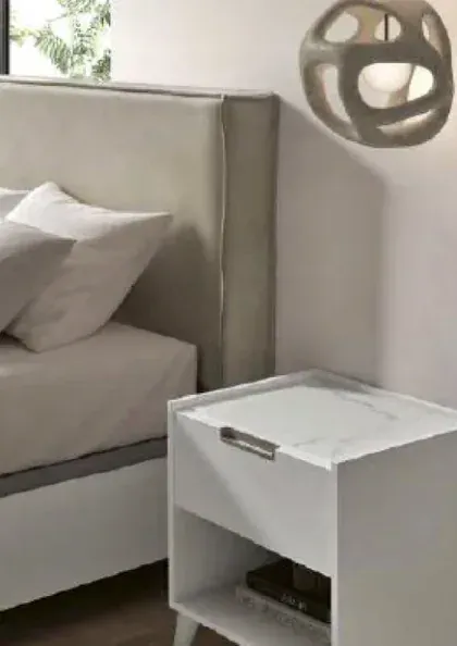 Rincón de dormitorio gris con una cama, una almohada blanca y una pequeña mesita de noche blanca bajo una lámpara de pared.