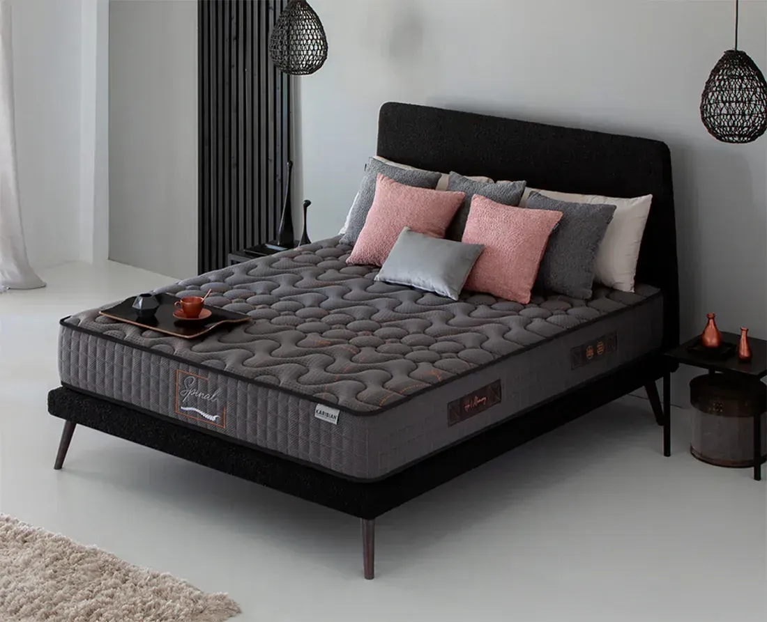 Dormitorio moderno con cama tapizada en gris, colchón blanco, almohadas negras y una manta oscura.