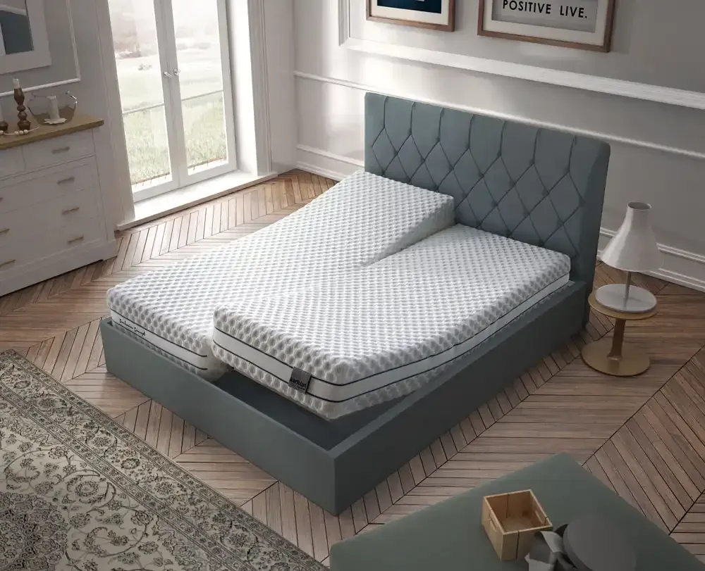 Cama tapizada en gris en un dormitorio luminoso con una gran ventana, mesita de noche y alfombra estampada.