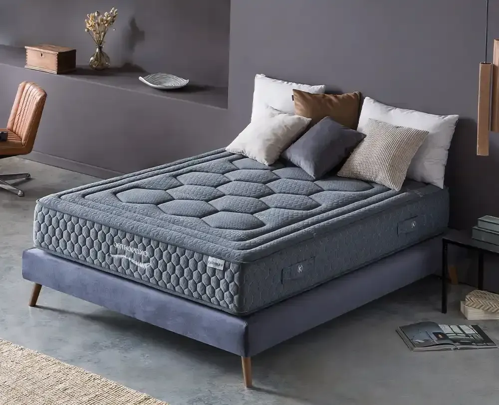 Dormitorio moderno con cama tapizada en gris, colchón blanco, almohadas negras y una manta oscura.