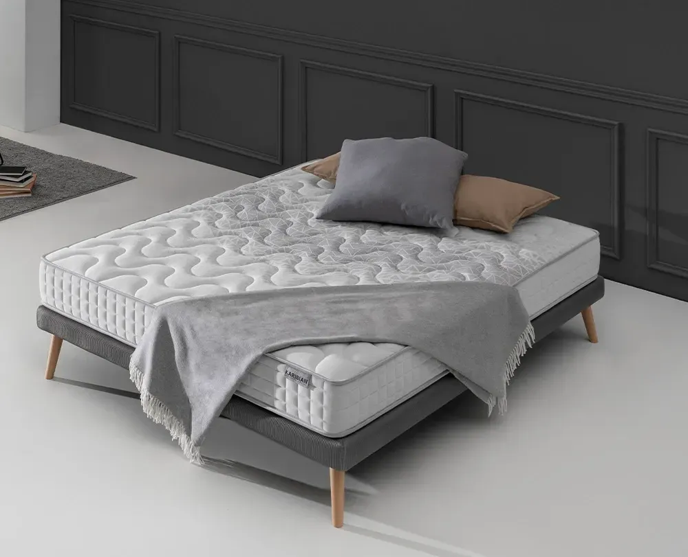 Dormitorio moderno con cama tapizada en gris, colchón blanco, almohadas negras y una manta oscura.