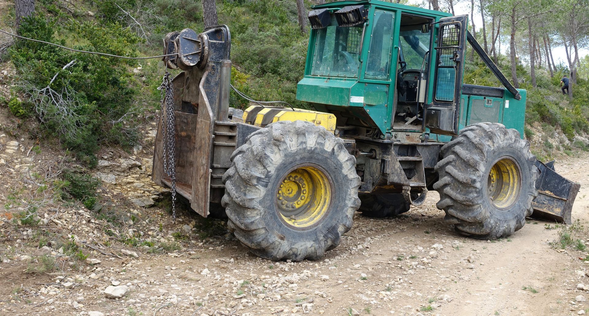 Tracteur forestier vert sur un chemin de terre dans une zone boisée.