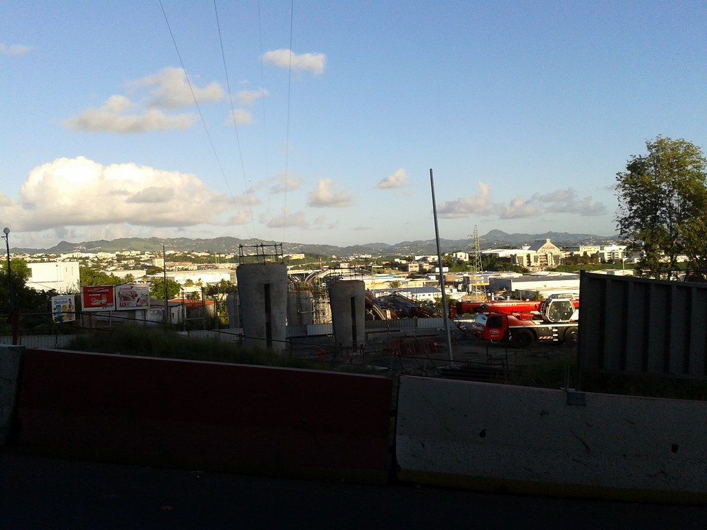 Chantier de construction avec piliers en béton gris, engins orange et paysage urbain sous un ciel nuageux.