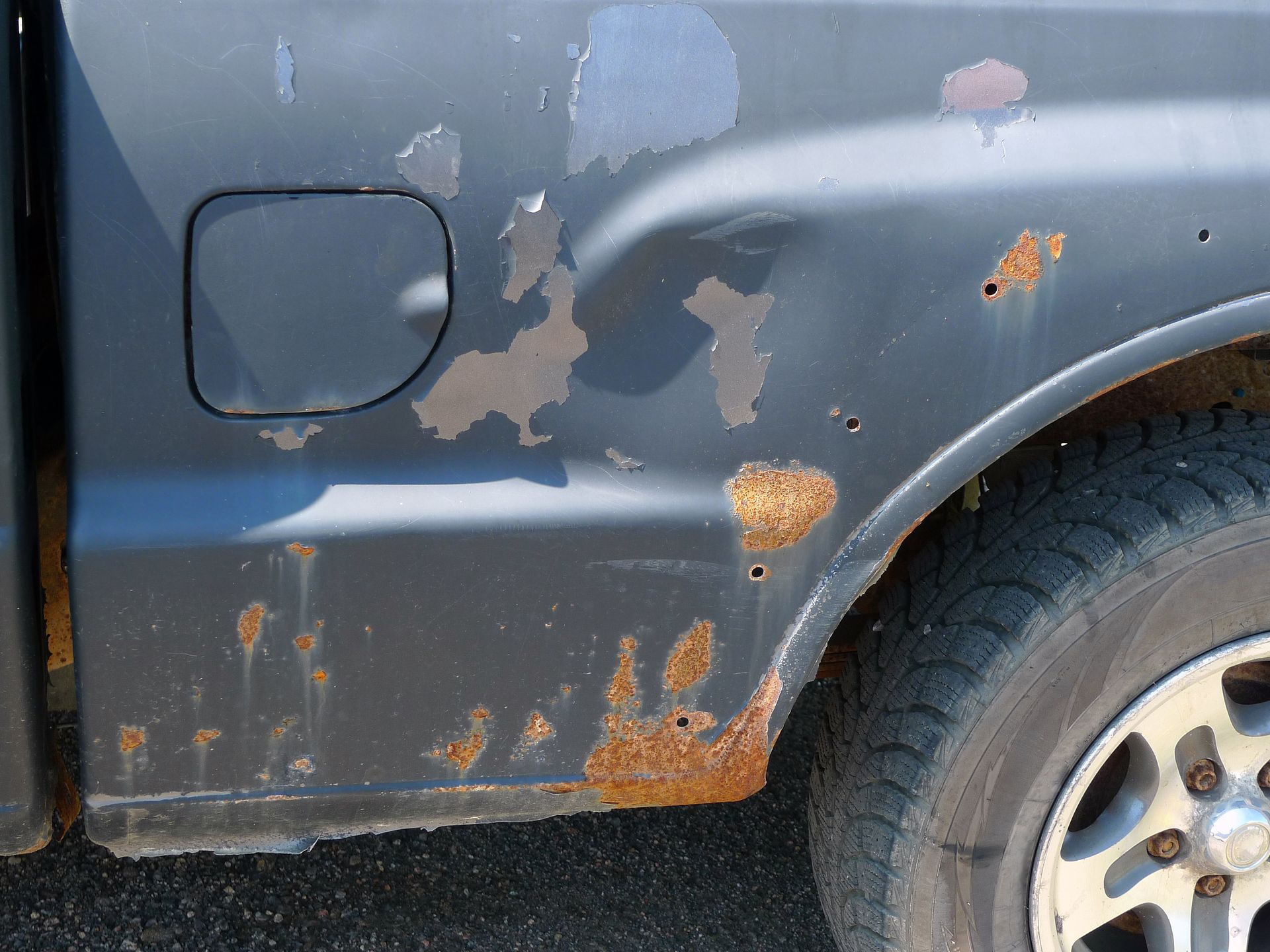 Benne de camion noire fortement rouillée, peinture écaillée, présentant des dommages près du pneu.