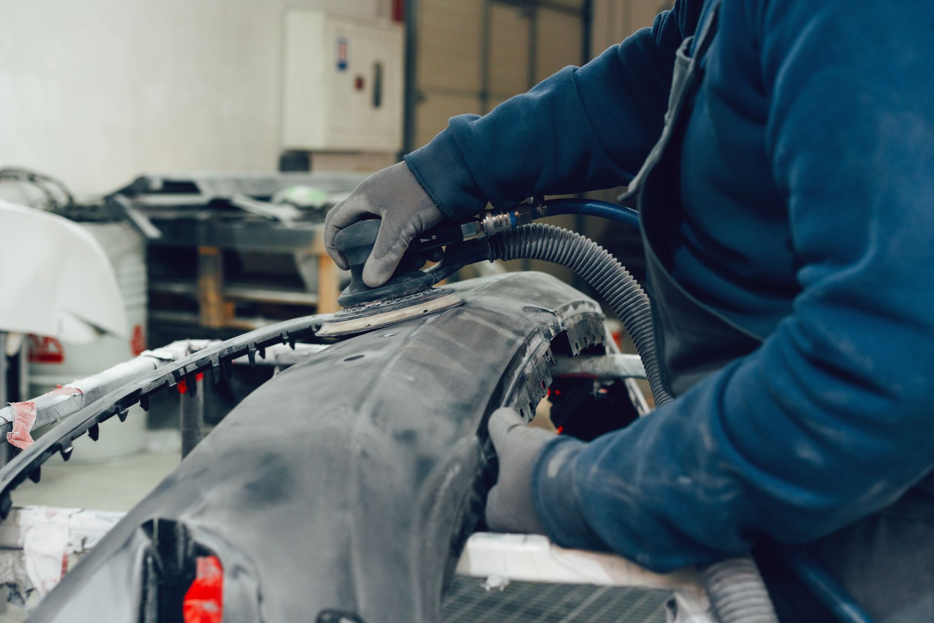 Une personne utilise une ponceuse sur un pare-chocs de voiture dans un atelier, portant des gants et une chemise bleue à manches longues.