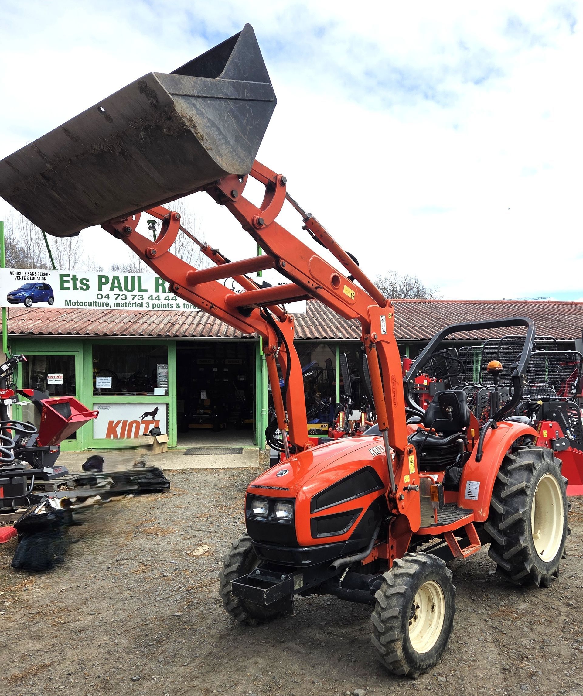 Tracteur orange avec godet de chargeur frontal relevé, stationné devant un magasin de matériel agricole.