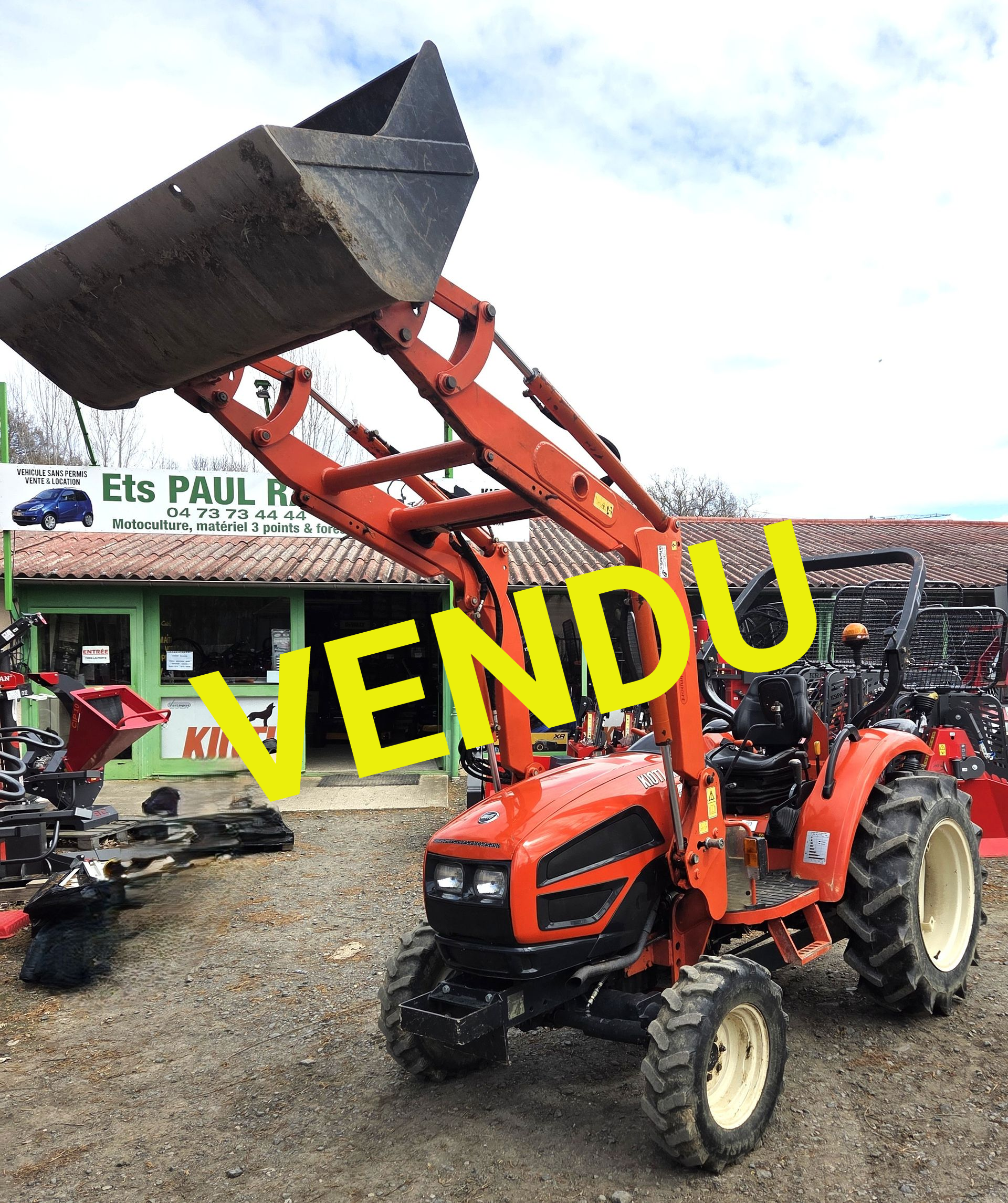 Tracteur orange avec godet de chargeur frontal relevé, stationné devant un magasin de matériel agricole.