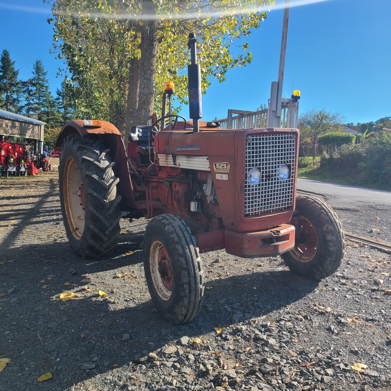 Un tracteur rouge est garé sur un terrain gravillonné près d'une clôture et d'une petite voiture jaune-bleue.