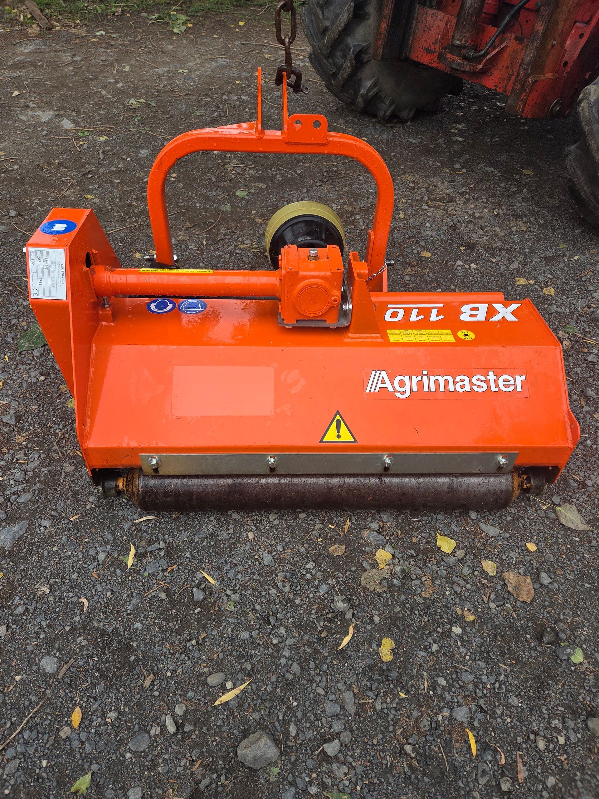 Accessoire de tracteur en métal rouge avec chaînes, à l'extérieur sur l'herbe, à côté de palettes empilées.