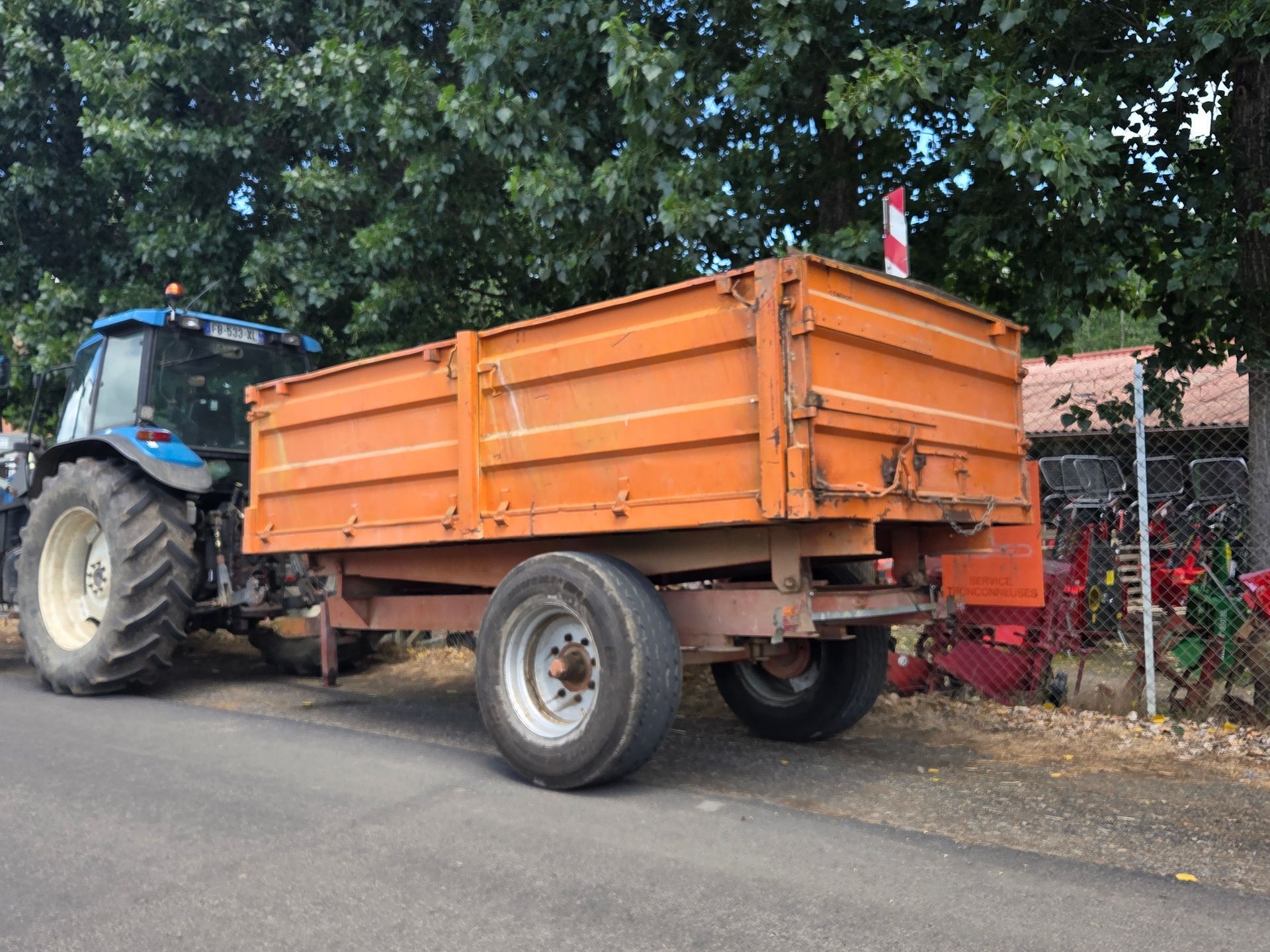 Une remorque agricole rouge avec une grande pancarte rouge « VENDU » garée dans une rue ensoleillée.