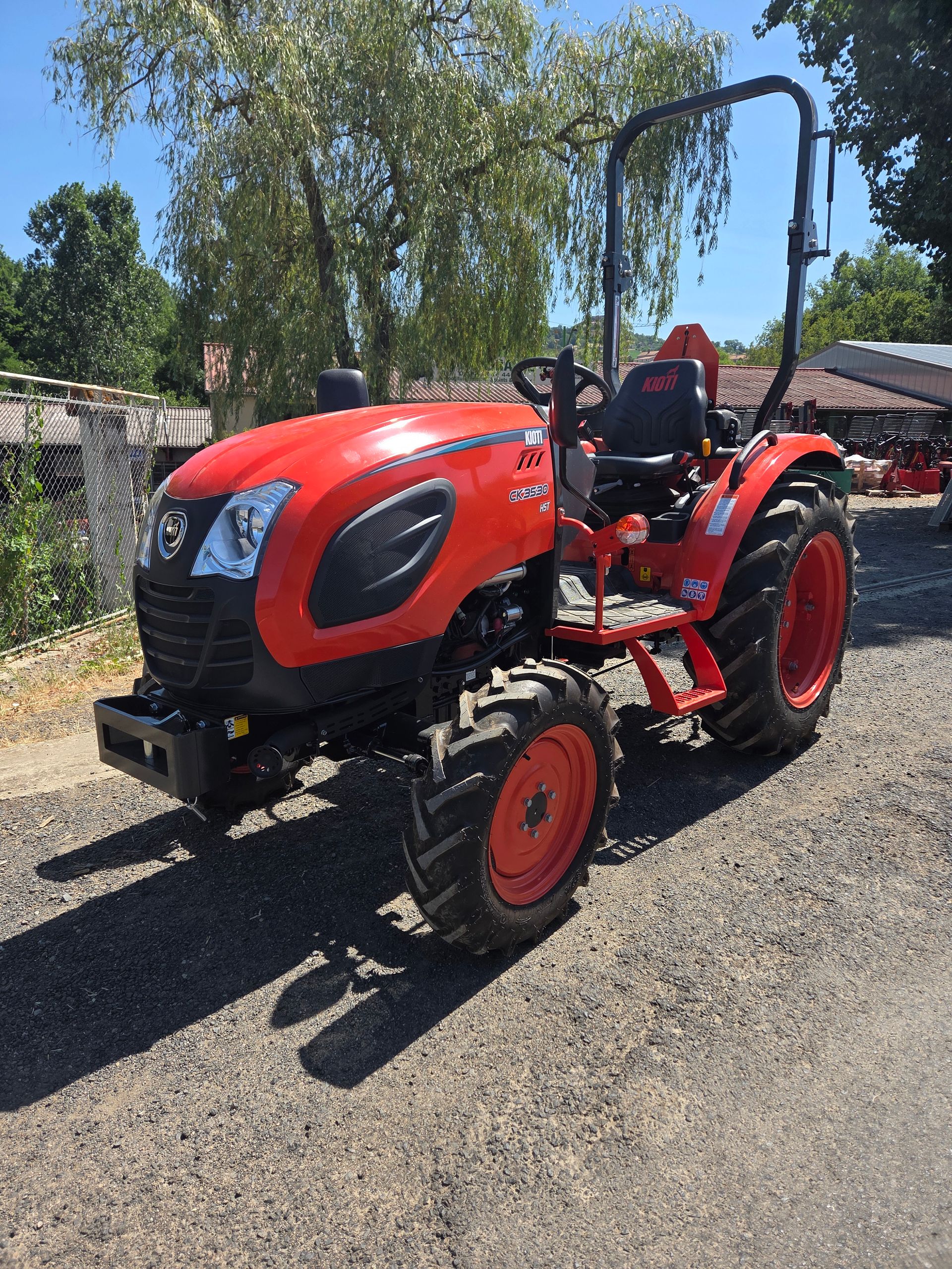 Tracteur orange garé sur l'herbe, à côté d'une clôture et d'arbres, sous un ciel bleu.