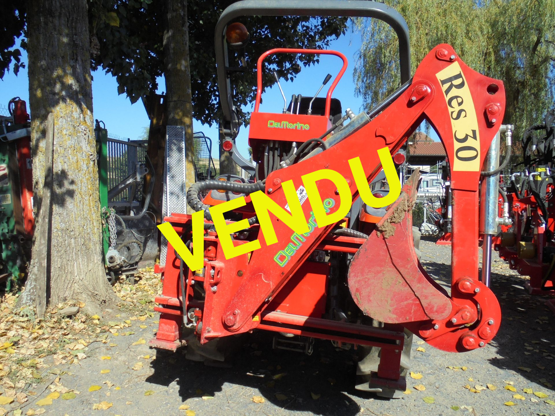 Une chargeuse-pelleteuse rouge est garée à l'extérieur, parmi les arbres et les feuilles mortes.
