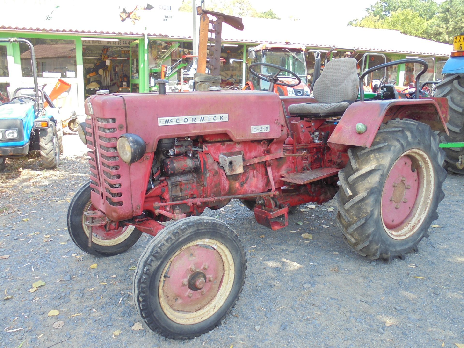 Tracteur rouge ancien garé sur du gravier, avec un tracteur bleu derrière, sous des arbres.